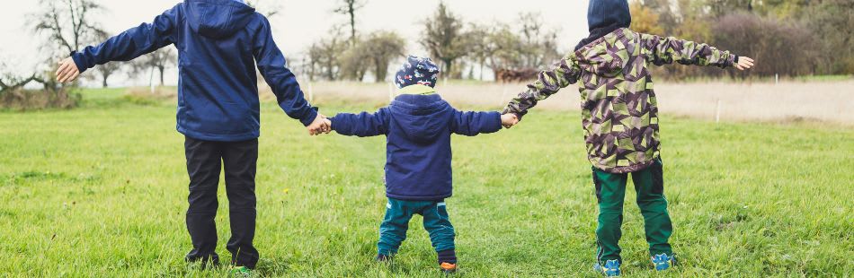 Camera behind three children dressed in jackets and hats walking hand in hand on some grass