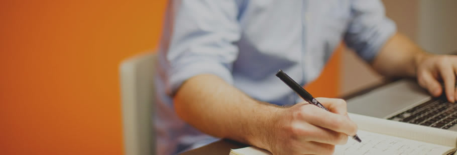 Man writing notes at a desk, with pen and paper and computer keyboard.