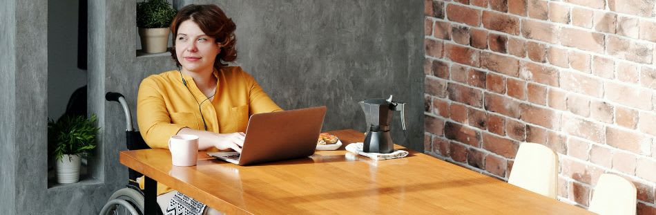 Woman in wheelchair sat at table looking off to the left with laptop and coffee
